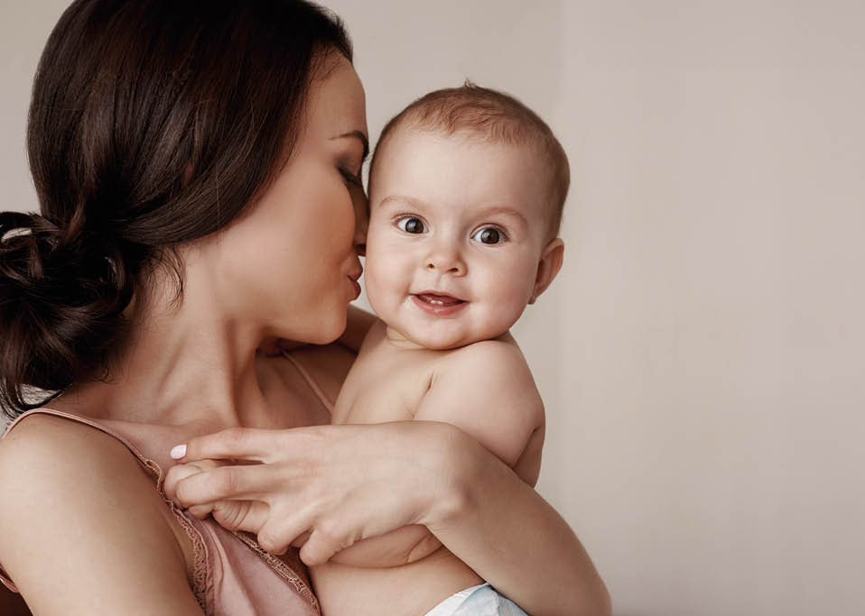 Young tender happy mother hugging her newborn baby smiling sitting on bed in morning. Copy space.