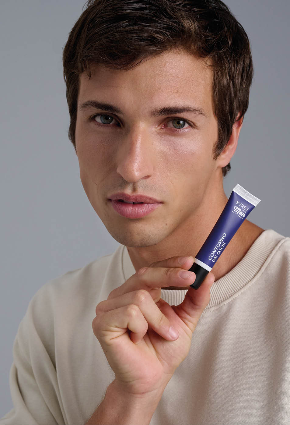 Persona con cabello corto y oscuro que sostiene cerca del rostro un tubo peque o de producto cosm tico con dise o azul y blanco mientras mira hacia adelante con expresi n neutra. Descripci n generada con IA