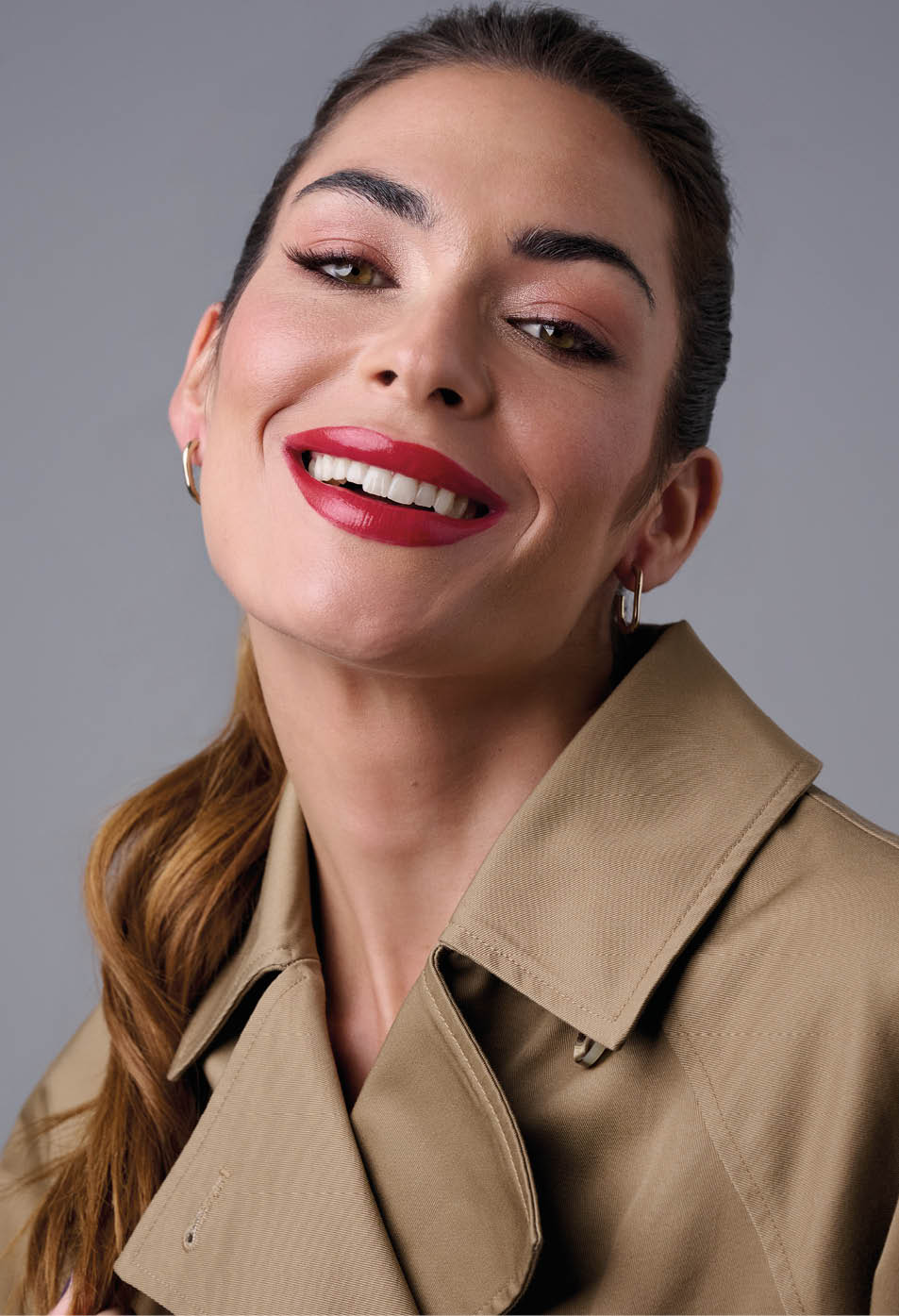 Persona adulta con cabello recogido y labios pintados de rojo sonriendo mientras lleva una gabardina beige. Descripci n generada con IA