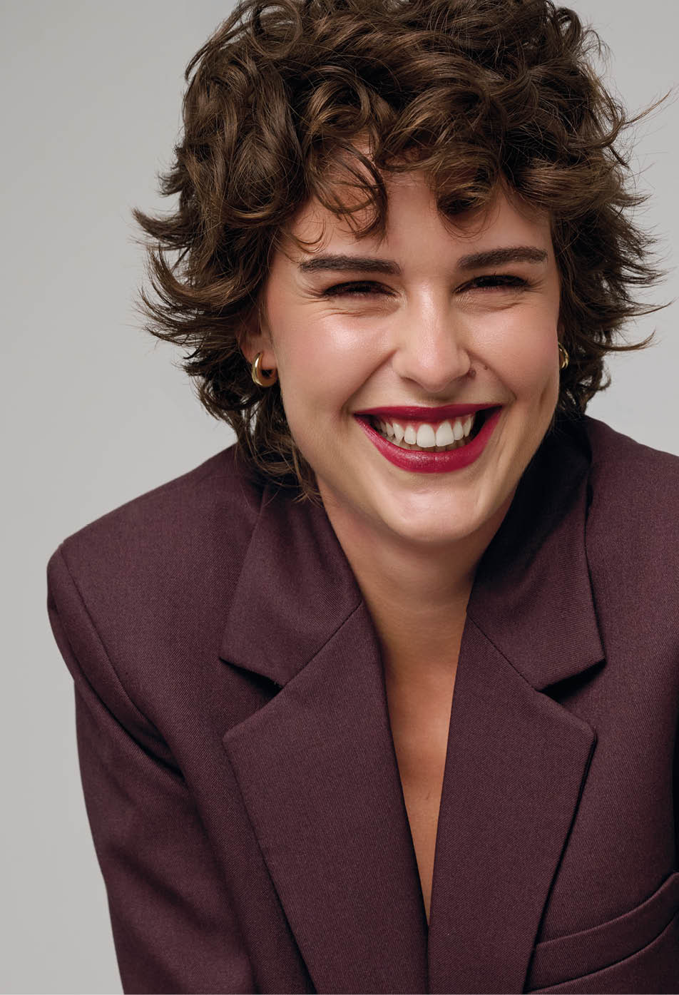 Persona con cabello rizado y corto que sonr e ampliamente mientras lleva una chaqueta de color vino. Descripci n generada con IA
