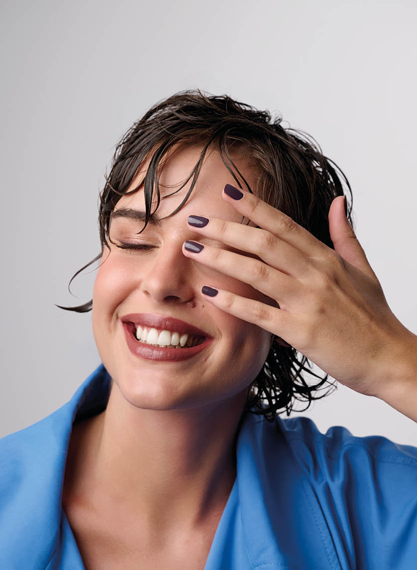 Persona con cabello corto y mojado sonriendo con los ojos cerrados mientras se cubre parcialmente el rostro con una mano que tiene las u as pintadas de color oscuro y viste una prenda azul. Descripci n generada con IA