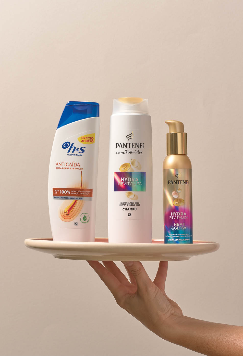 Una mano sostiene un plato con tres botellas de productos para el cuidado del cabello de las marcas Ohs y Pantene sobre un fondo neutro. Descripci n generada con IA