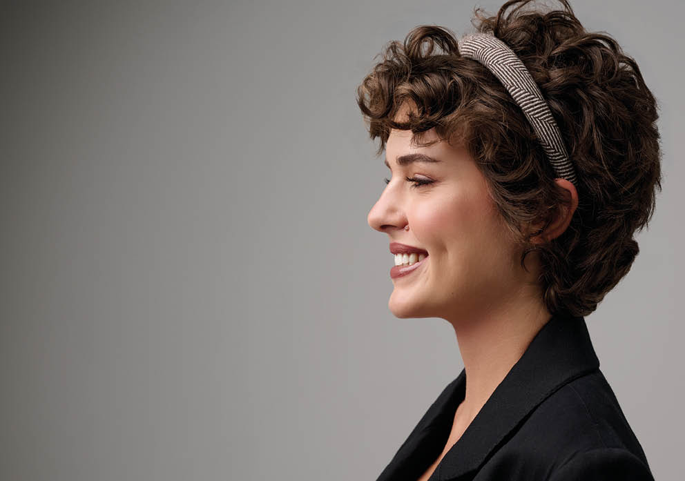 Persona con cabello rizado corto y una diadema sonriente de perfil con chaqueta oscura sobre un fondo gris. Descripci n generada con IA