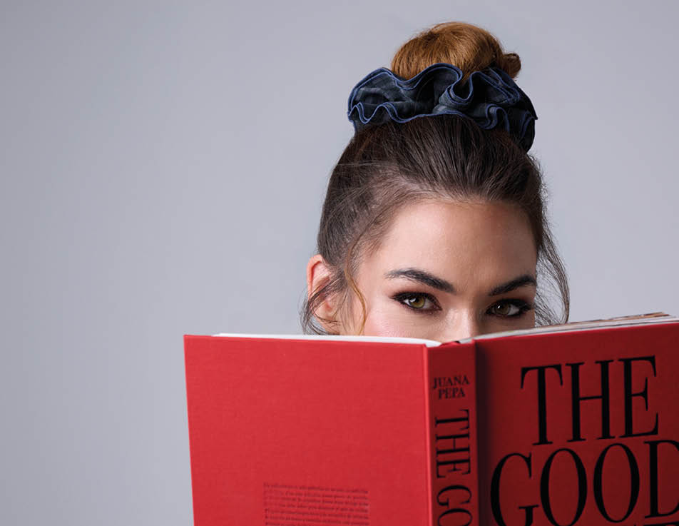 Una persona con el cabello recogido en un mo o alto y una goma negra sostiene un libro rojo frente al rostro mientras mira hacia adelante. Descripci n generada con IA