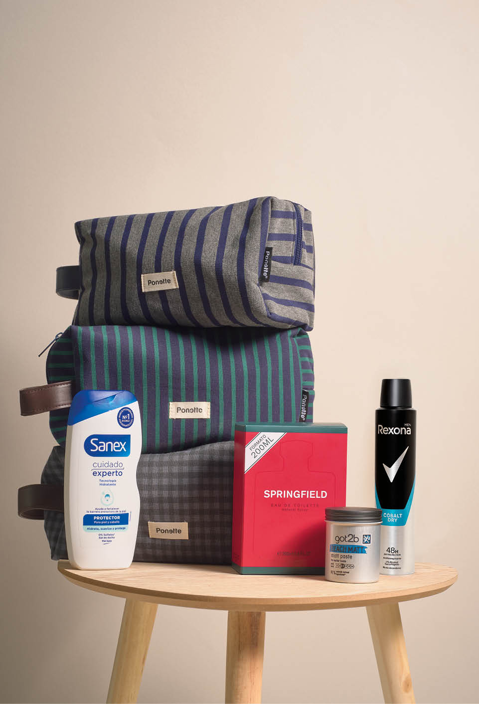 Tres neceseres de tela con rayas en tonos azules y verdes apilados sobre una mesa de madera junto a productos de cuidado personal como desodorante, crema para el cabello y gel de ducha. Descripci n generada con IA