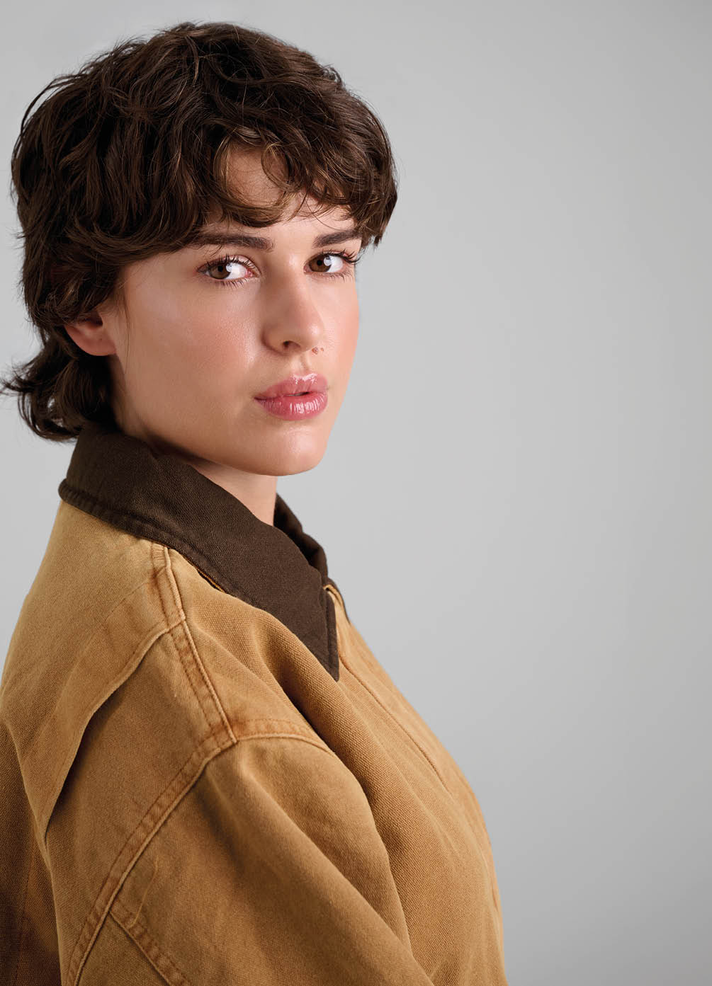 Persona con cabello corto y rizado que lleva una chaqueta marr n claro con cuello oscuro mirando hacia la c mara con expresi n neutral sobre un fondo gris claro. Descripci n generada con IA