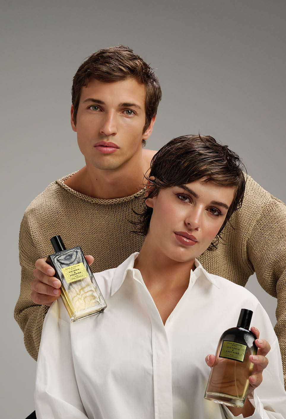 Dos adultos con cabello corto posan juntos sosteniendo frascos de perfume mientras visten ropa casual en un fondo neutro. Descripci n generada con IA