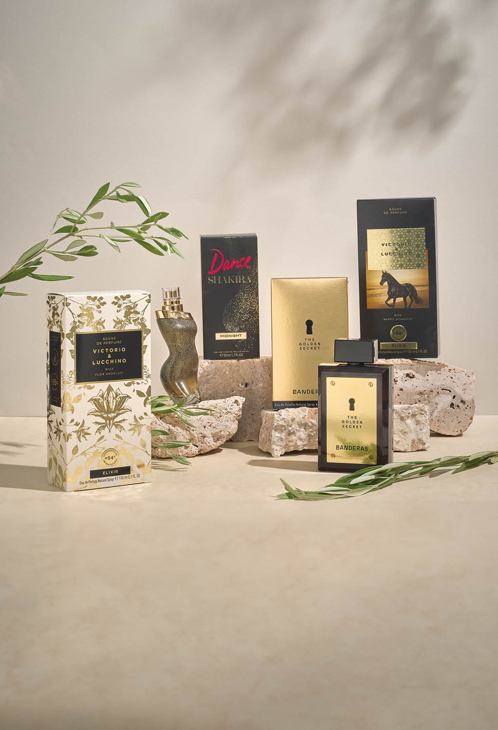 Varios frascos y cajas de perfumes decorativos est n dispuestos sobre una superficie clara con ramas verdes y piedras como elementos decorativos en un ambiente iluminado suavemente. Descripci n generada con IA