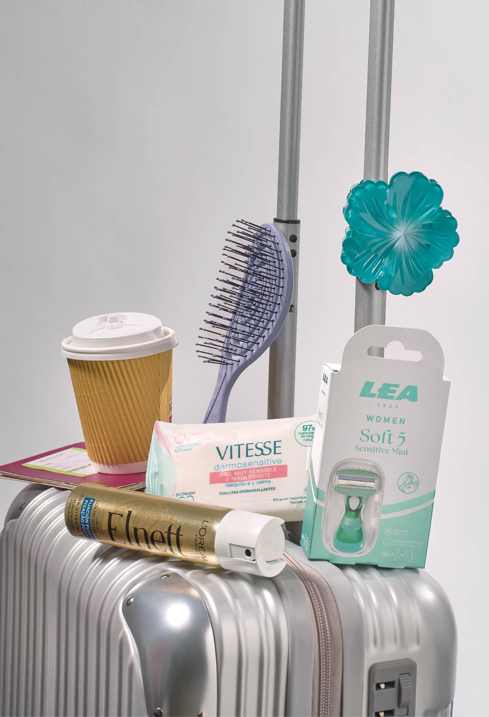Maleta plateada con asa extendida que sostiene varios objetos personales como un vaso de caf , un cepillo, toallitas h medas, un envase de cera para el cabello y un paquete de depilaci n. Descripci n generada con IA